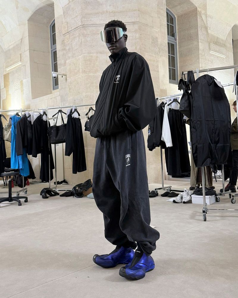 An adult male model with dark skin poses in a black tracksuit and visor sunglasses inside an event space with racks of clothing.
