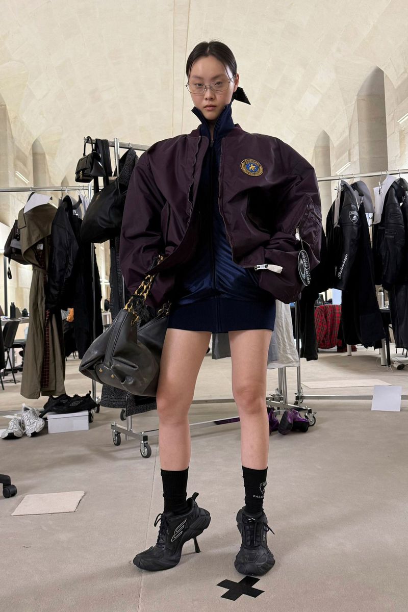 Stylish woman in a jacket, dress, socks and high heel shoes poses in a studio with clothing racks and minimalist decor. 