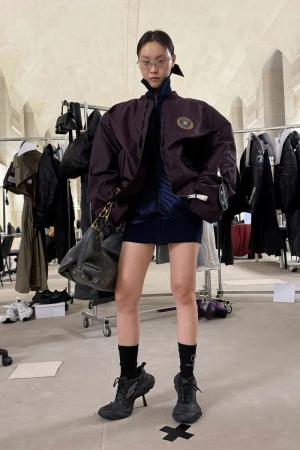 Stylish woman in a jacket, dress, socks and high heel shoes poses in a studio with clothing racks and minimalist decor. 