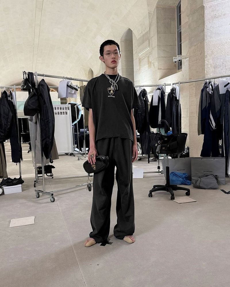Young adult poses in black casual wear with pendant jewelry indoors, backstage with clothing racks in the background.