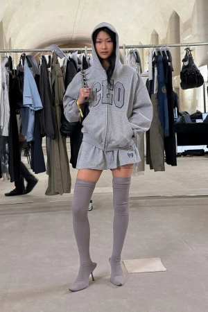 A young Asian woman models a gray hoodie and skirt with matching thigh-high socks and stiletto heels in a light studio setting.