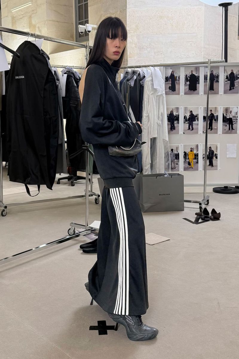 A slender Asian woman in a black tracksuit and heels stands in a light, neutral setting with clothing racks and fashion images.