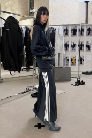 A slender Asian woman in a black tracksuit and heels stands in a light, neutral setting with clothing racks and fashion images.