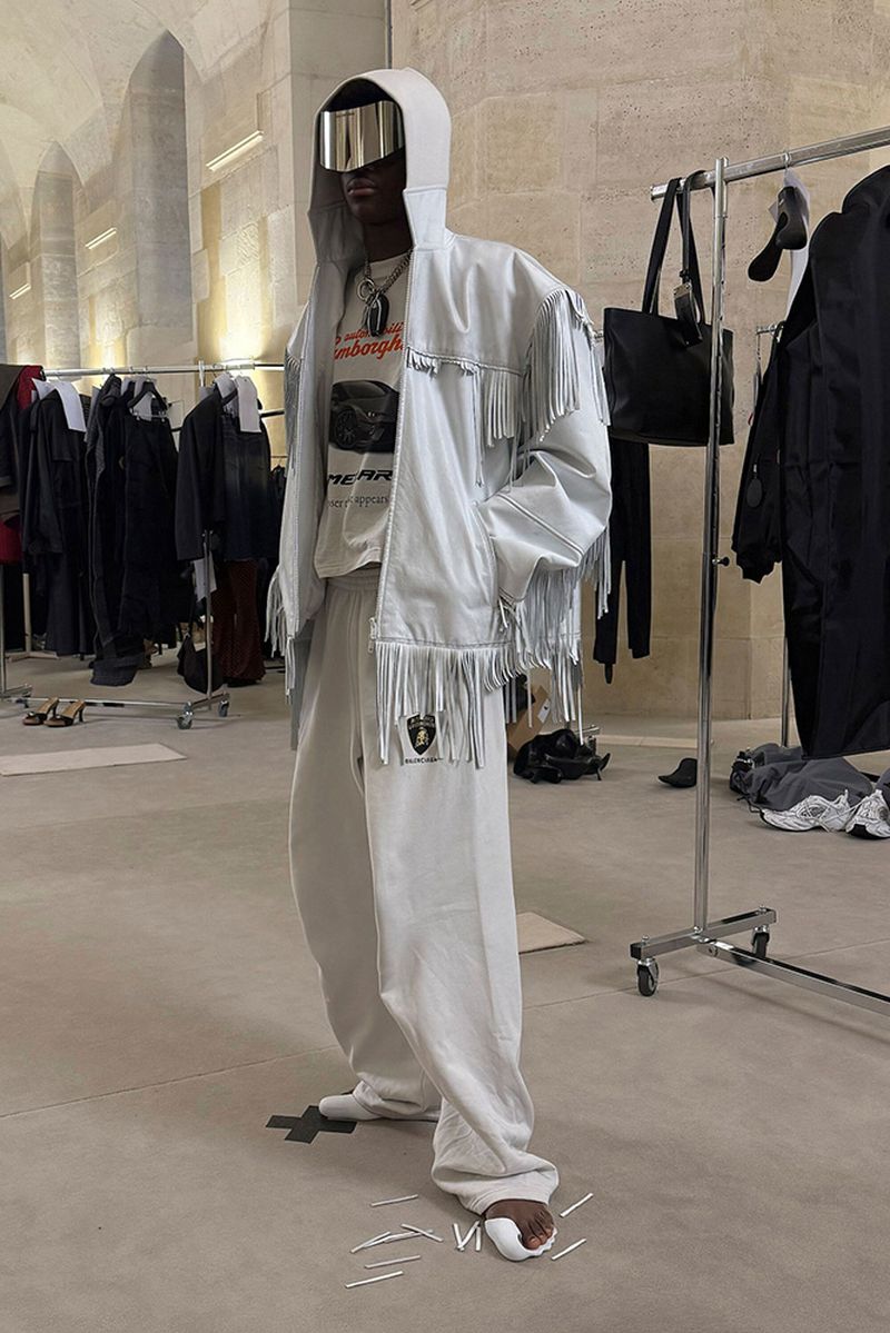 An adult male in a hooded jacket with fringe and silver visor stands indoors in a light-toned outfit, showcasing street-style fashion.