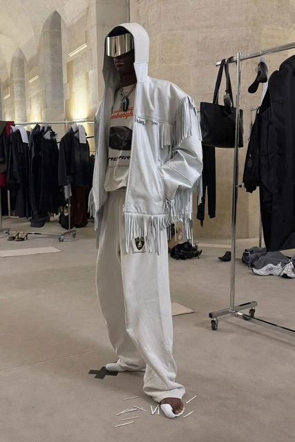 An adult male in a hooded jacket with fringe and silver visor stands indoors in a light-toned outfit, showcasing street-style fashion.