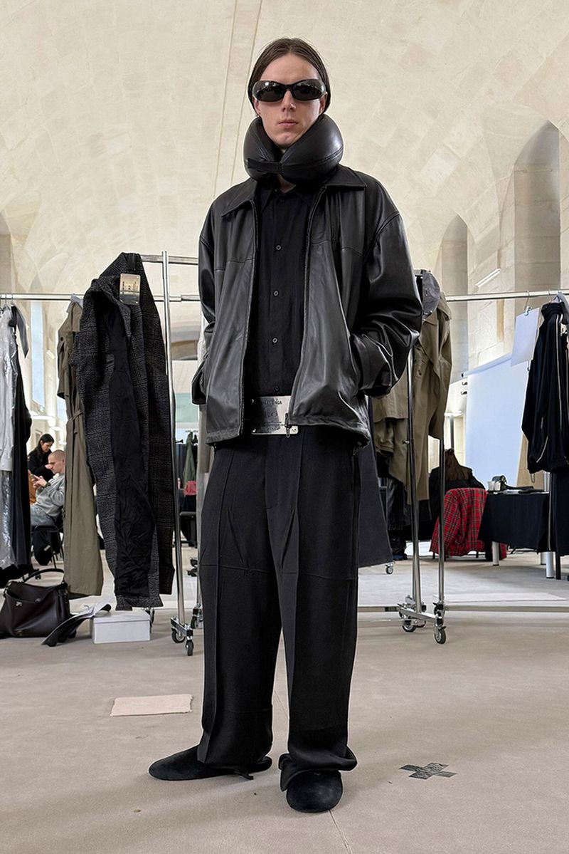 Full-length portrait of a male model in all-black avant-garde fashion, featuring a padded collar, leather jacket, and wide-leg pants.