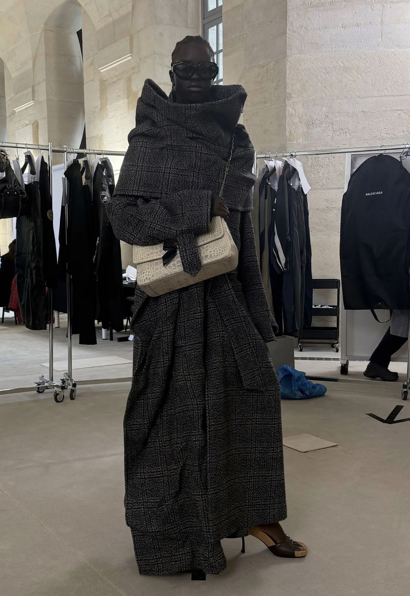 Editorial fashion shot: African female model in oversized grey plaid coat, sunglasses, beige handbag, heels in backstage setting.
