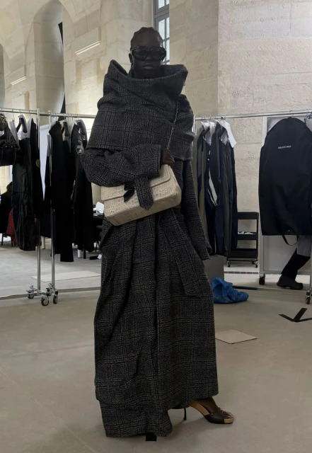 Editorial fashion shot: African female model in oversized grey plaid coat, sunglasses, beige handbag, heels in backstage setting.