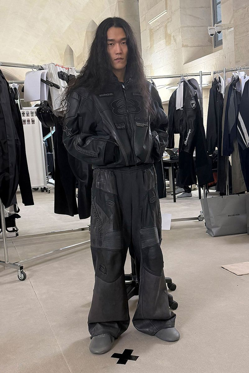 Adult Asian man in a black leather jacket and oversized pants posing indoors; wardrobe racks with clothes are in the background.
