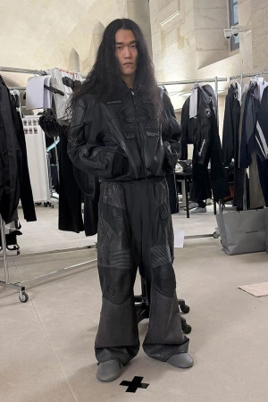 Adult Asian man in a black leather jacket and oversized pants posing indoors; wardrobe racks with clothes are in the background.