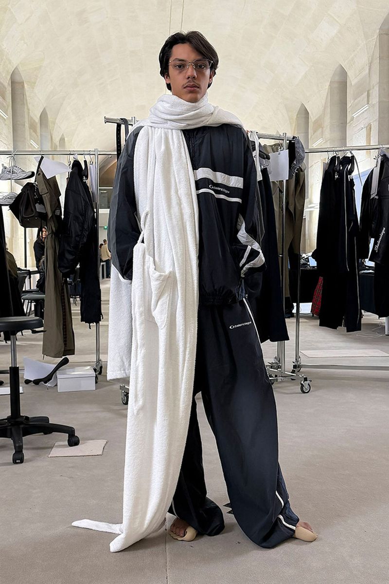 Standing adult in relaxed pose in an indoor setting, wearing a black and white tracksuit and white scarf, with clothing racks in the background.