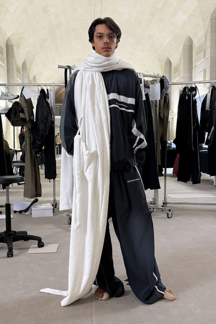 Standing adult in relaxed pose in an indoor setting, wearing a black and white tracksuit and white scarf, with clothing racks in the background.