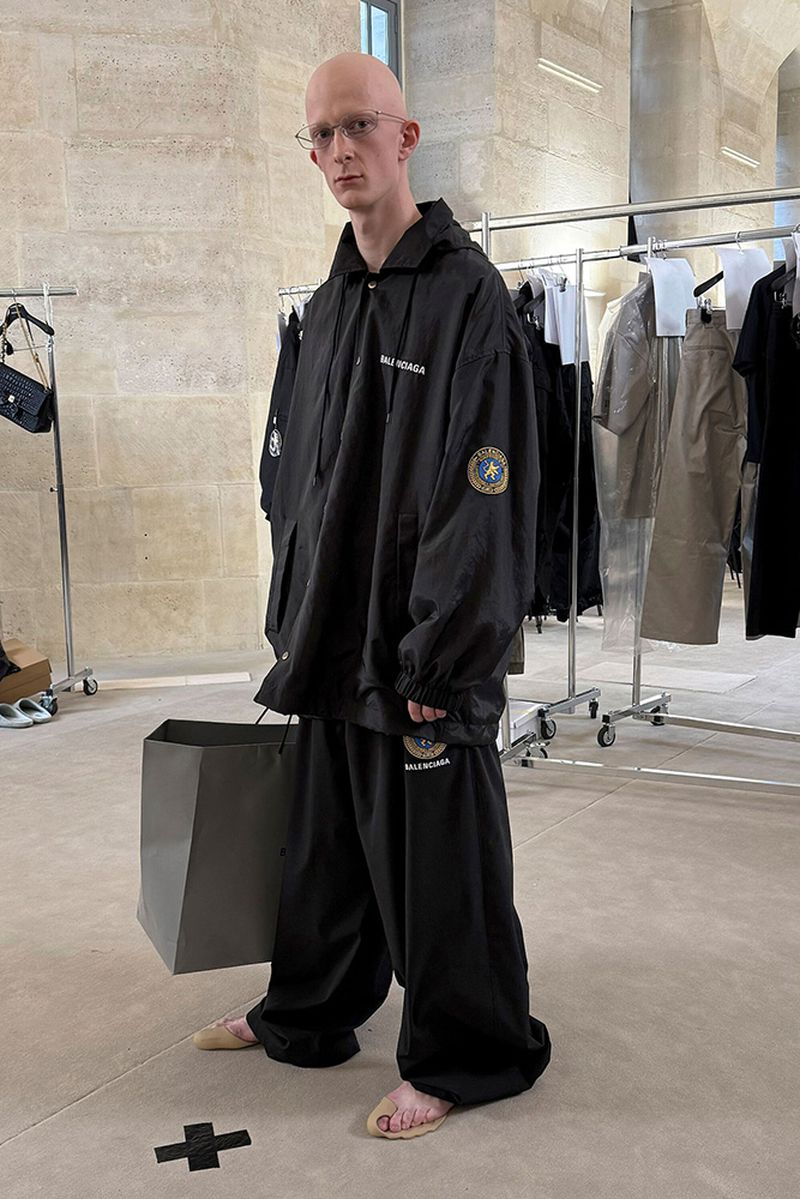 A bald adult man in a black Balenciaga tracksuit stands indoors with a shopping bag, near a rack of clothes in a fashion setting.