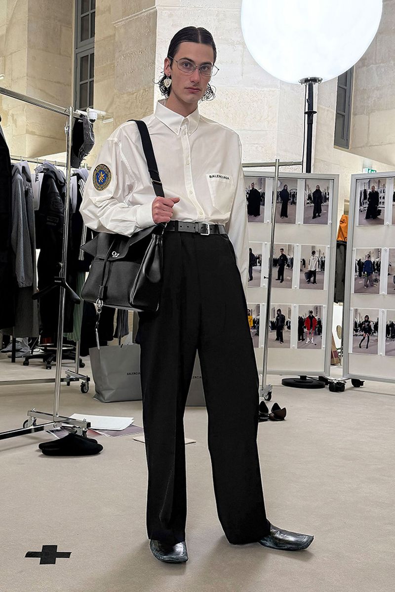 Male fashion model in button-down shirt, wide-leg pants, and silver shoes, standing indoors with a black shoulder bag.