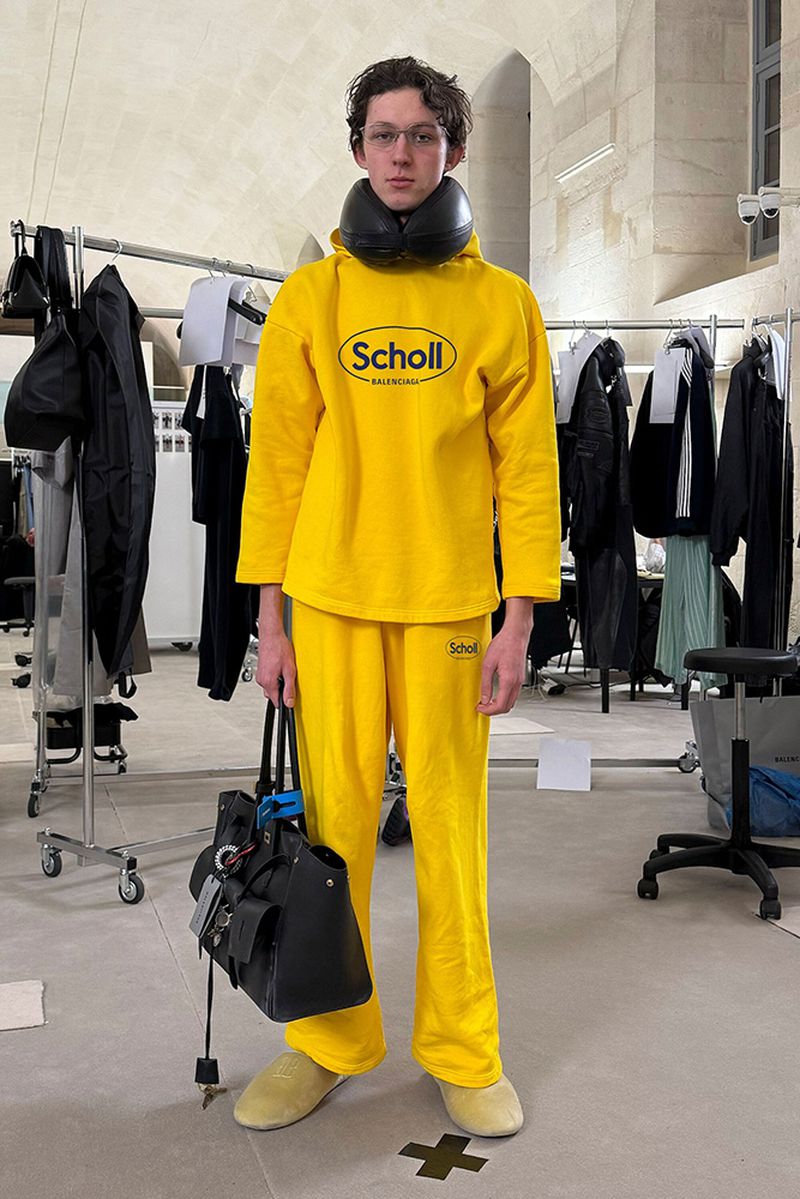 A young adult stands in a yellow Scholl brand outfit, holding a black bag, indoors with clothing racks in the background.