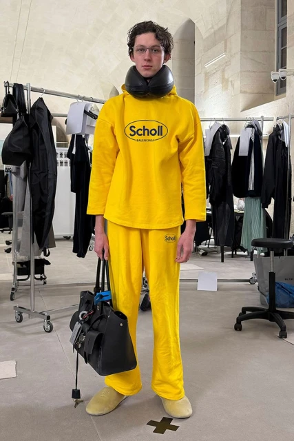 A young adult stands in a yellow Scholl brand outfit, holding a black bag, indoors with clothing racks in the background.