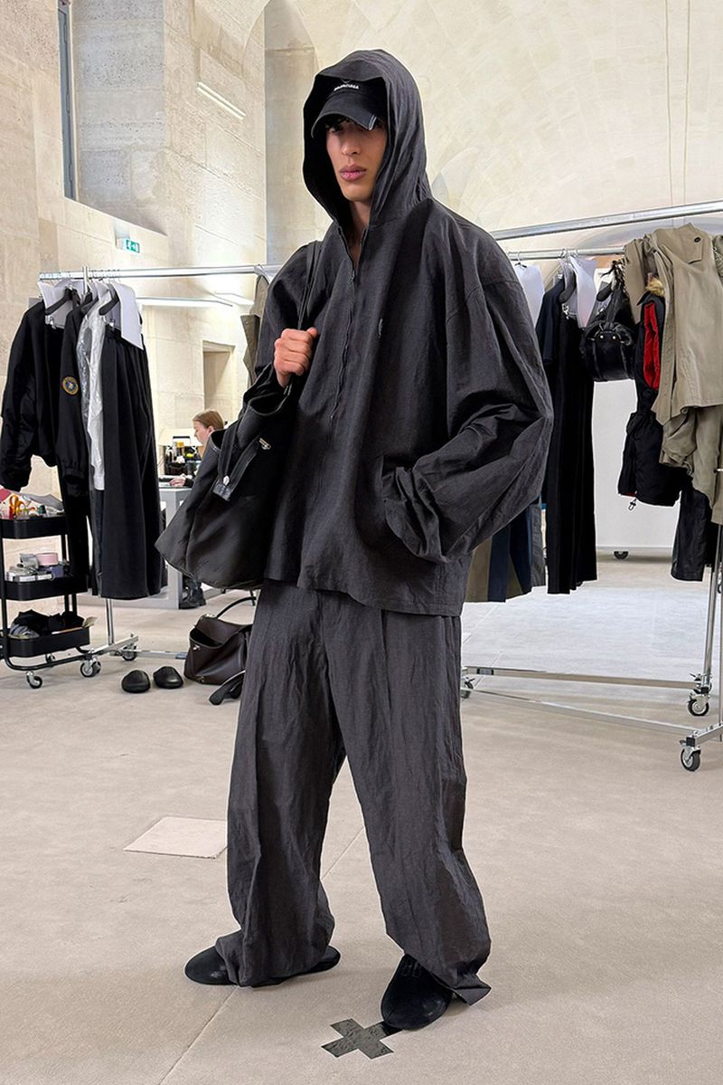 A slender adult male model stands in a stone-walled interior, wearing a dark gray hoodie, baggy pants, slides, and a baseball cap.