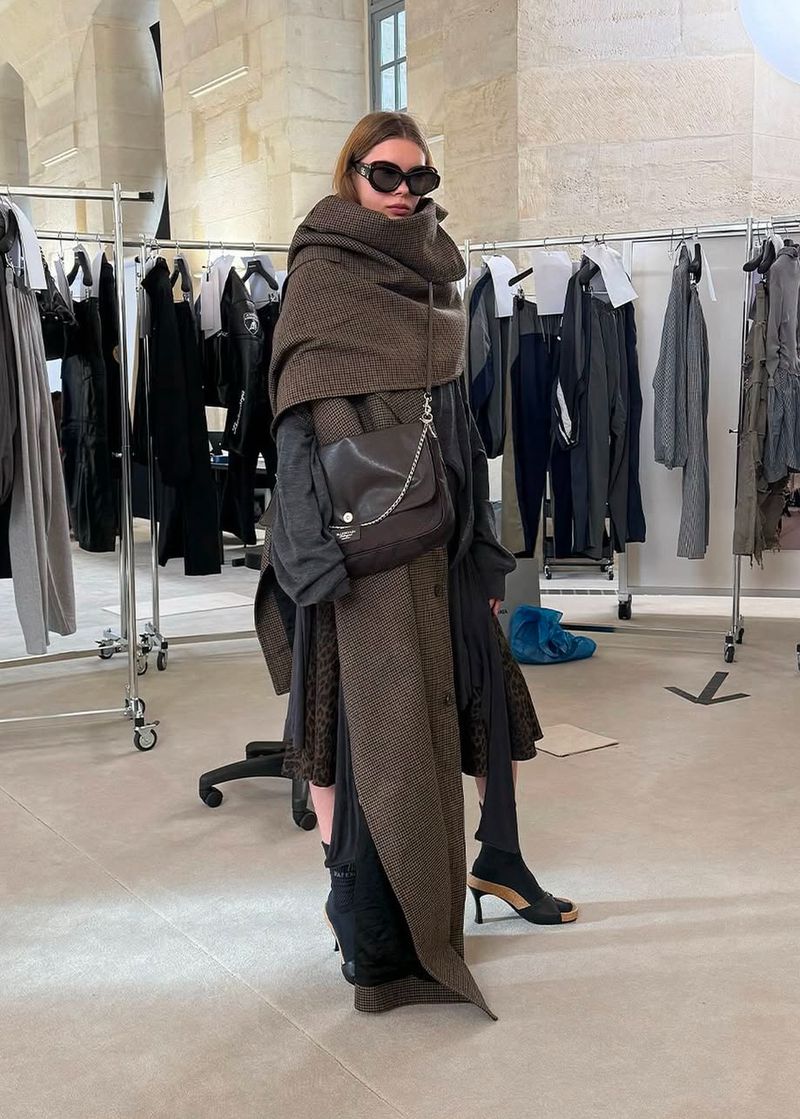 Woman wearing sunglasses and a brown patterned wrap and shoulder bag stands in an indoor area with clothing racks in the background.