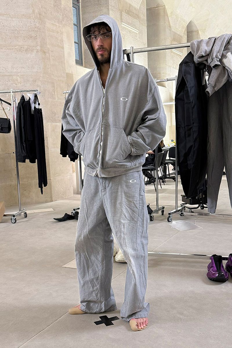 Man in casual grey sweatsuit with hoodie standing indoors in a naturally lit neutral space with clothing racks in the background.