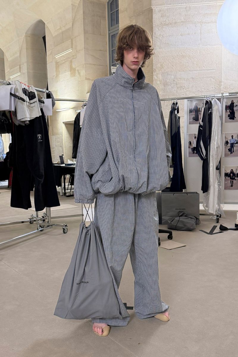 Young adult man in oversized grey checkered streetwear-style jacket and pants, holding a grey tote bag in an indoor location.