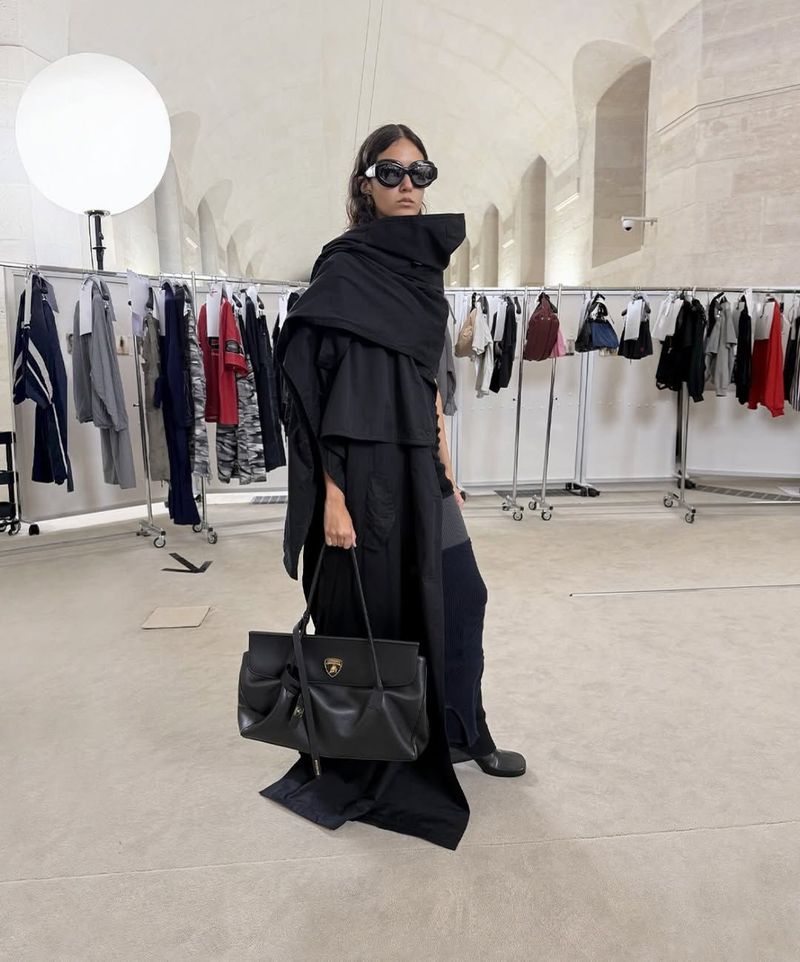 Fashionable woman wearing a draped black coat and sunglasses holds a black leather handbag in an indoor setting with clothing racks.