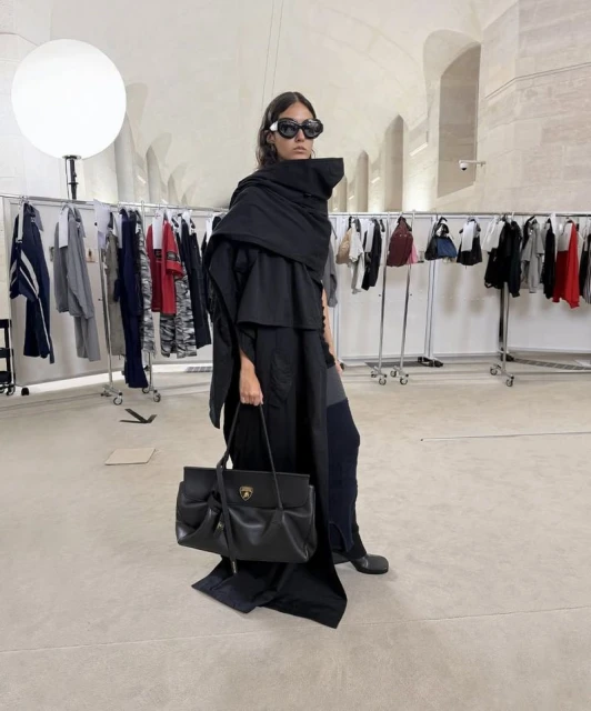 Fashionable woman wearing a draped black coat and sunglasses holds a black leather handbag in an indoor setting with clothing racks.