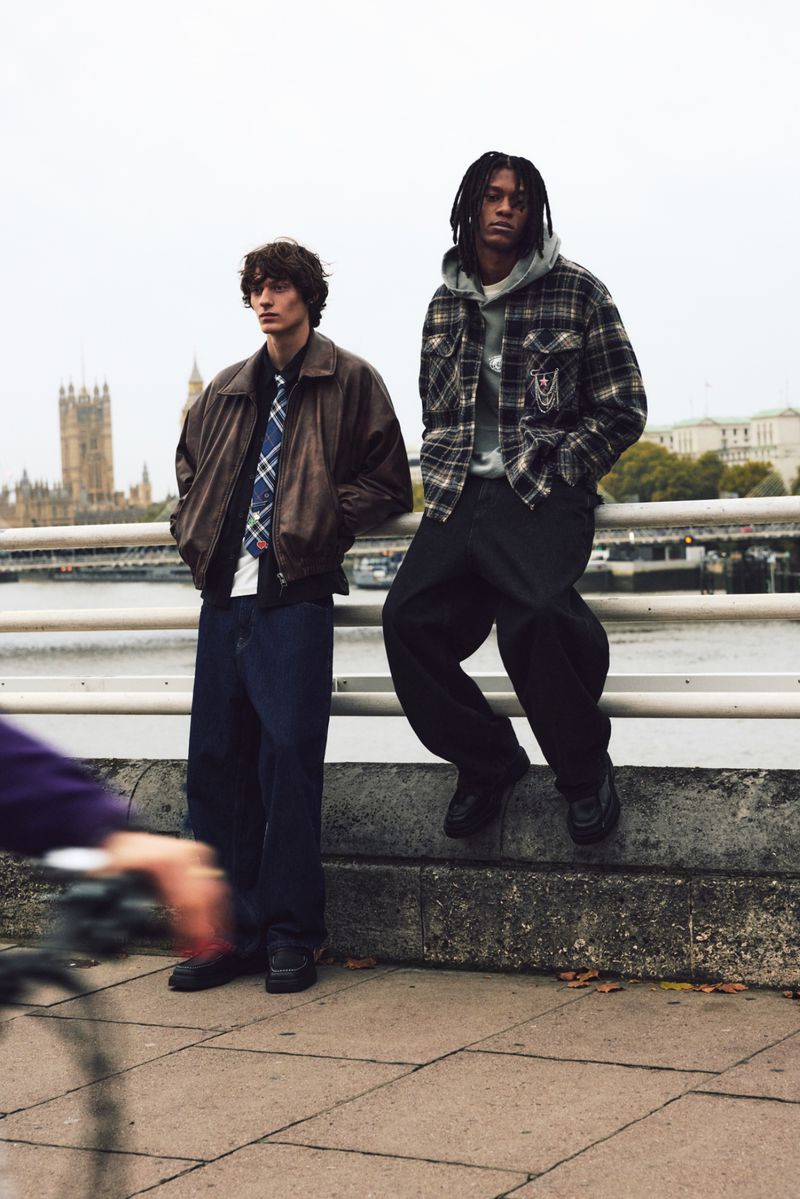 Two young adult men stand on a bridge in London, wearing fashionable jackets and trousers, with the Houses of Parliament in the background.