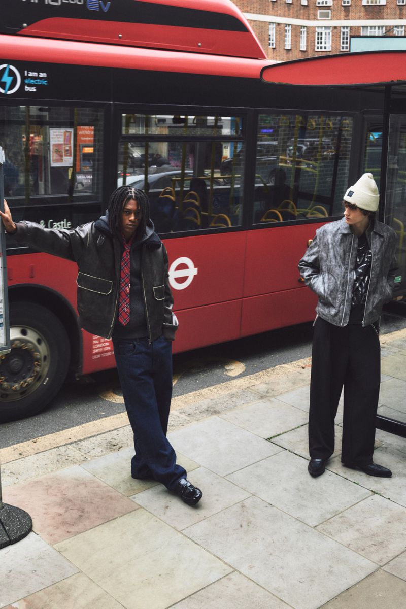 Two young adults pose in trendy streetwear next to a red double-decker bus on a city street with brick buildings in the background.