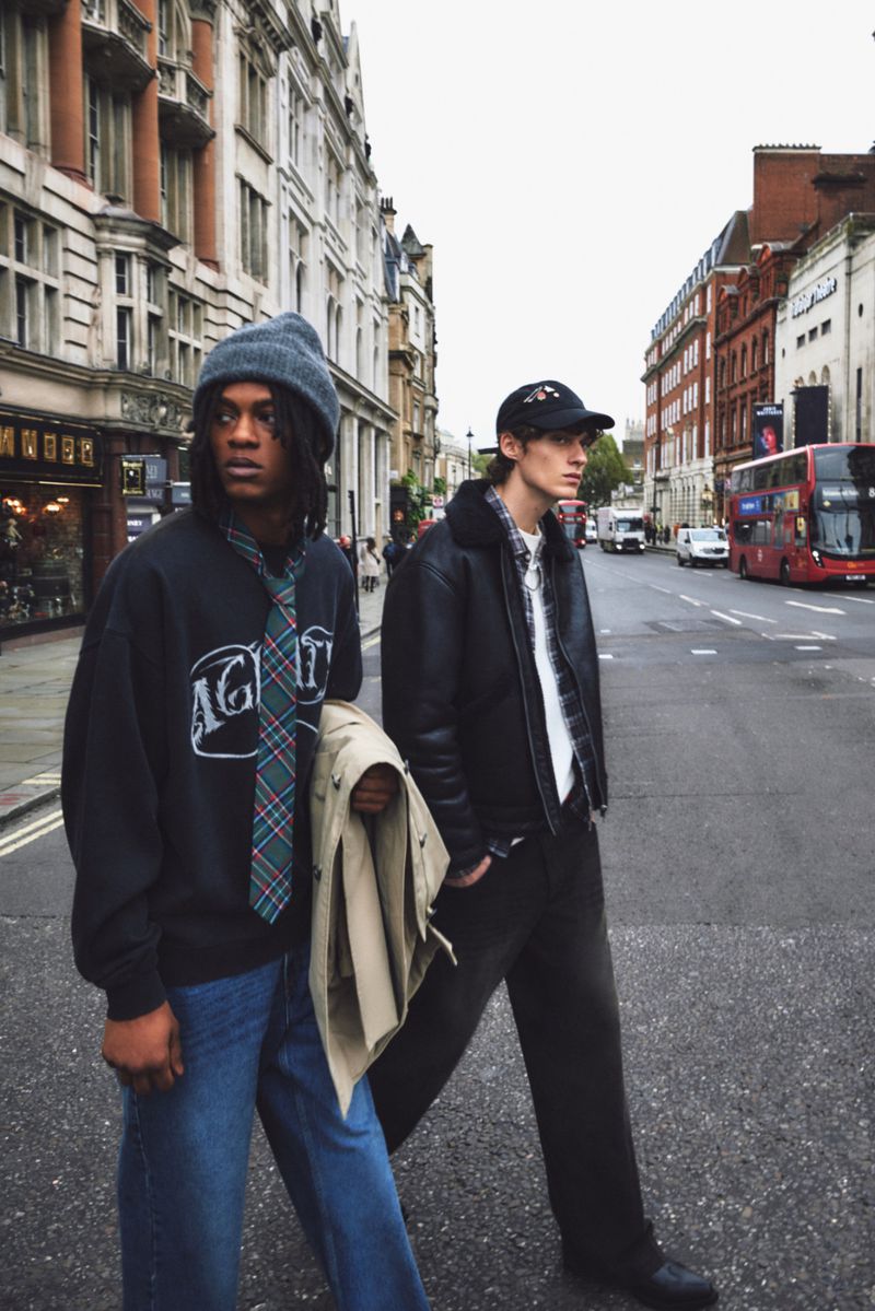 Two young adults pose on a city street in fashionable outfits, showcasing a blend of casual and stylish elements against an urban backdrop.