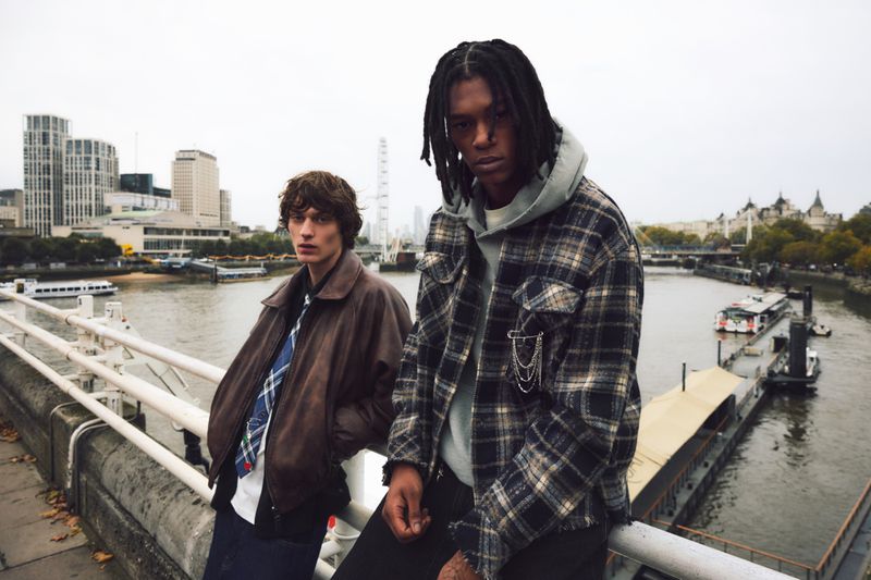 Two adult male models in outerwear stand on a bridge with a city and river in the background under an overcast sky, fashion editorial.