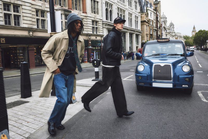 Two people cross a street with a blue London taxi cab in the background, set against a backdrop of urban buildings on a cloudy day.