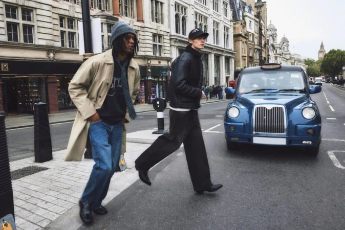 Two people cross a street with a blue London taxi cab in the background, set against a backdrop of urban buildings on a cloudy day.