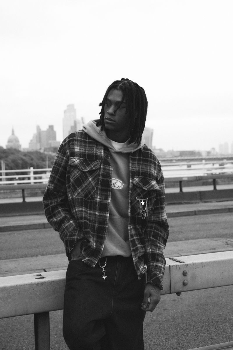 Black and white outdoor portrait of a Black male wearing a plaid jacket and hoodie, with a city skyline and cloudy sky.