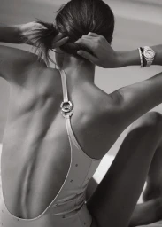 Model seen from behind wearing a light-coloured swimsuit with back straps and a white wristwatch, adjusting her hair.