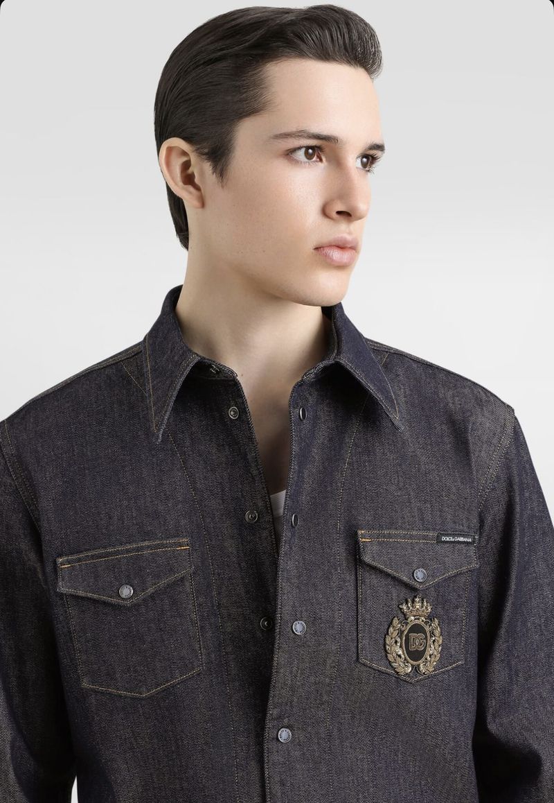 Close-up of a young adult male wearing a dark denim shirt with chest pockets and a decorative logo against a solid light-grey background.