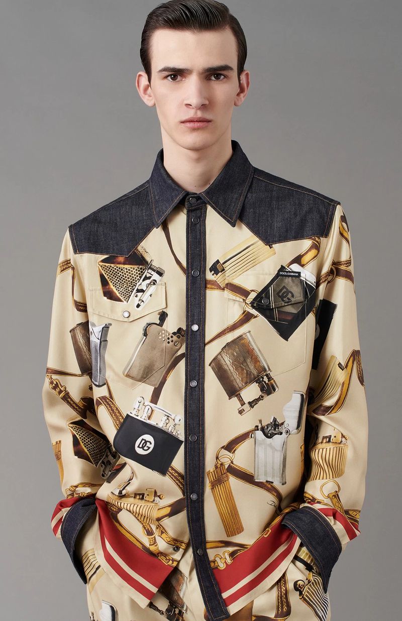 Male fashion model wears a patterned beige button-down shirt with denim shoulders in a studio shot against a neutral gray background.