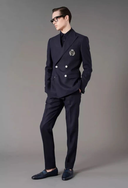 Full-length studio shot of an adult male fashion model wearing a dark blue double-breasted suit, collared shirt, black glasses, and loafers.