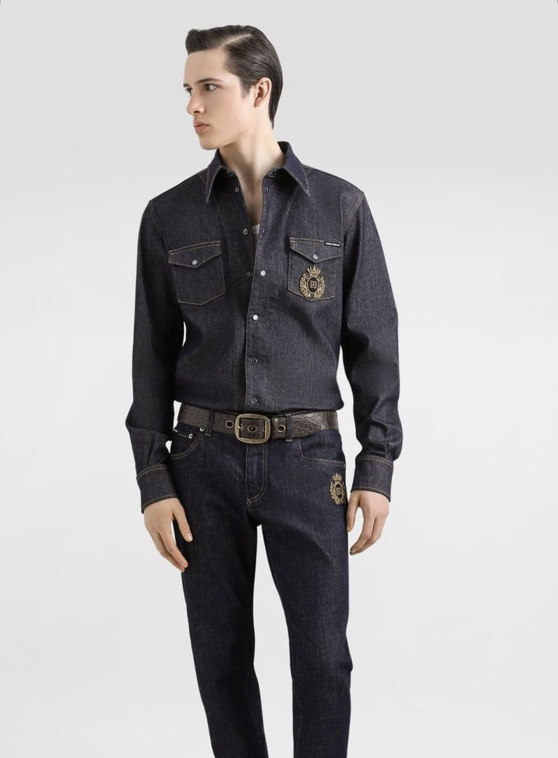 Male model wearing dark denim shirt and jeans with gold crest embroidery on a seamless white background.