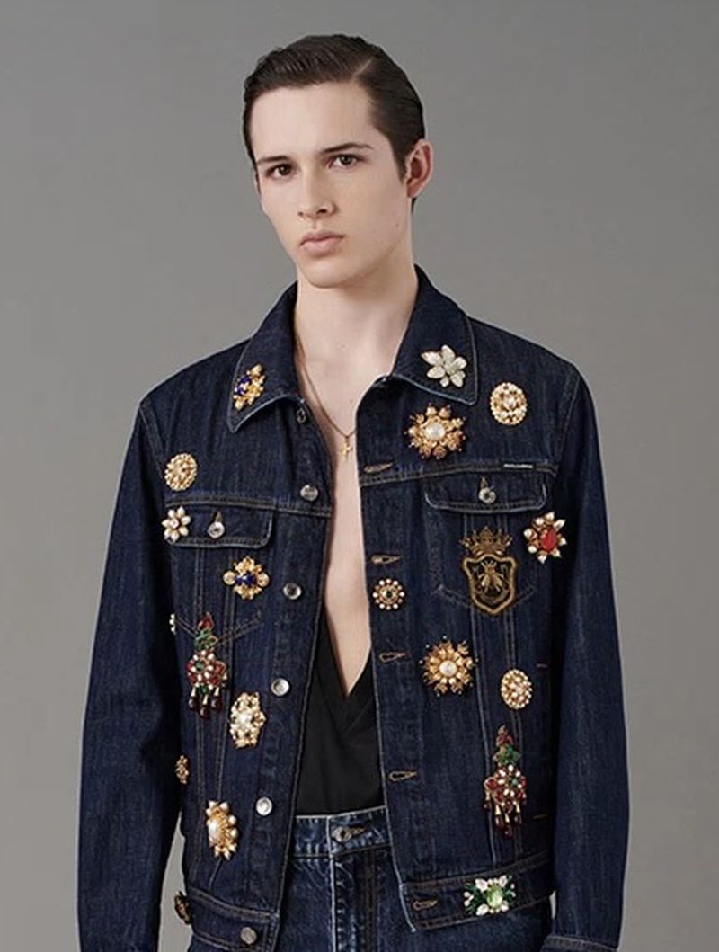 Male model wears a denim jacket with ornamental brooches in a studio shot. Fashionable and stylish clothing, neutral background.