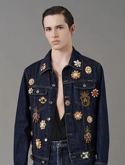 Male model wears a denim jacket with ornamental brooches in a studio shot. Fashionable and stylish clothing, neutral background.
