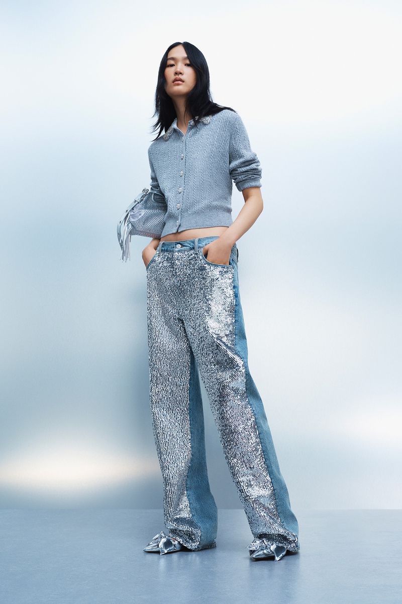 Fashion model posing in studio wearing a grey button-down sweater, denim and sequin pants, and silver shoes against a gradient background.
