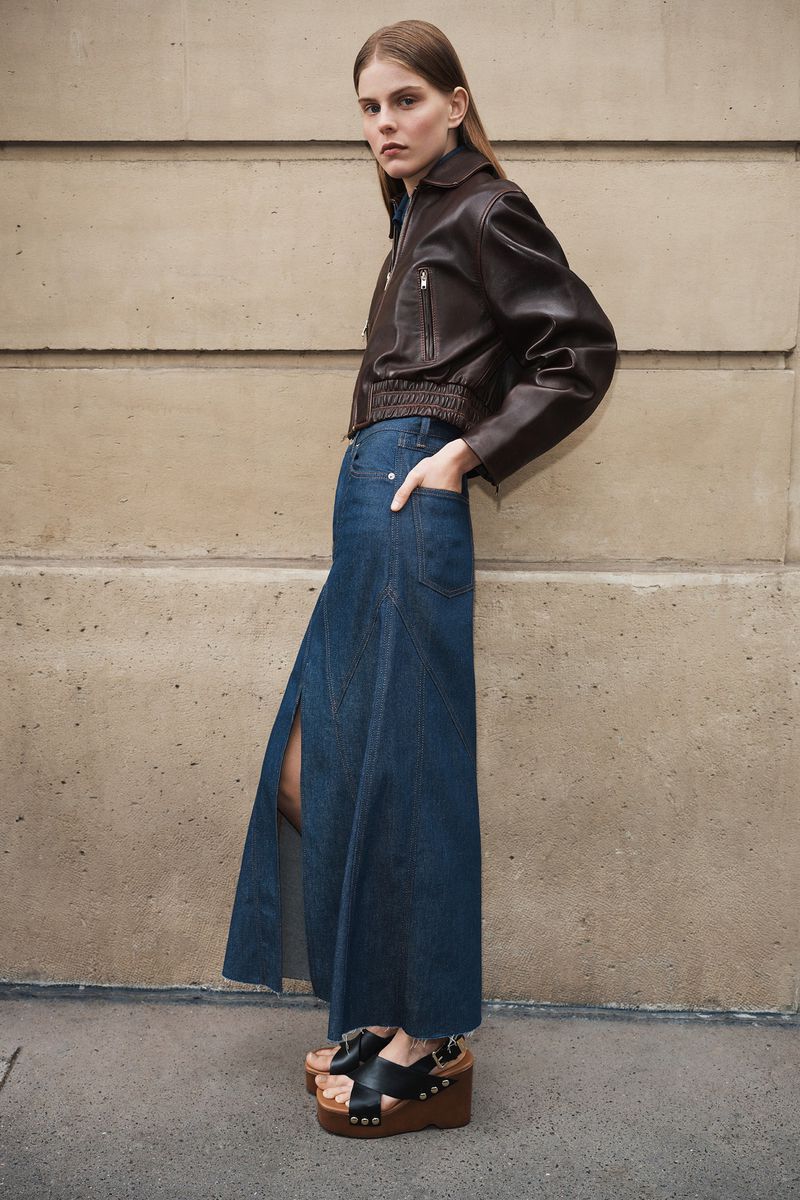Model in brown leather jacket and long denim skirt standing against a stone wall