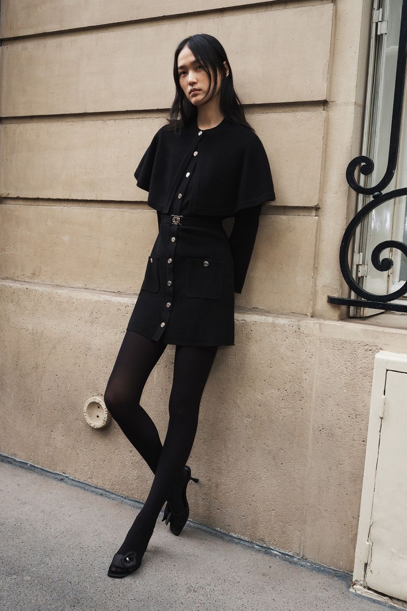 Asian woman in a short black dress with a cape and black stockings poses against a neutral-colored wall in an outdoor setting.