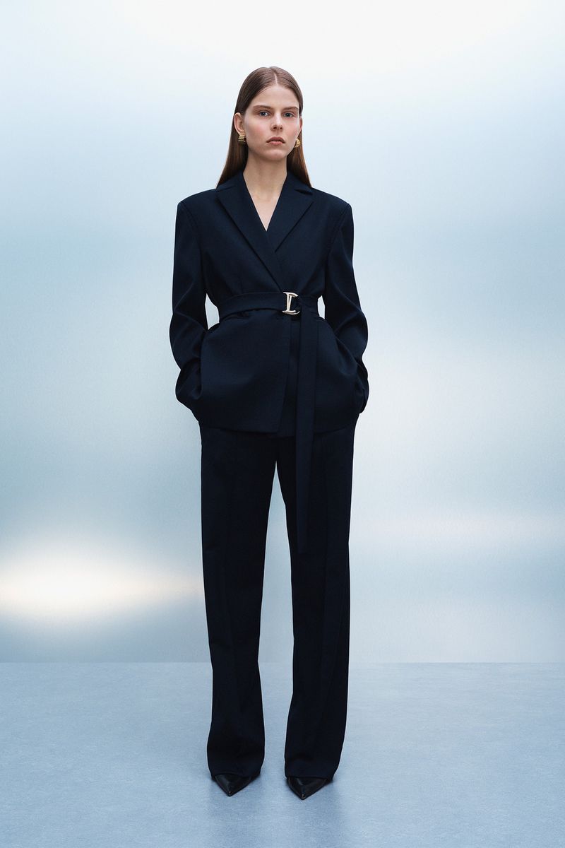 Elegant adult woman in a dark belted suit and heels poses against a light blue studio backdrop. Fashion-forward, minimalist style.