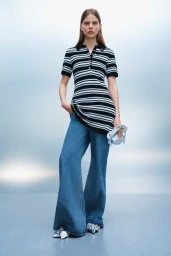 Model wearing a black and white striped polo top and wide-leg blue denim jeans against a gradient studio backdrop.