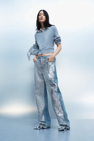 Model wearing a blue cardigan and sequined wide-leg jeans against a gradient blue studio backdrop for Maje.