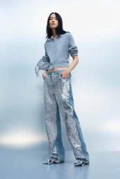 Model wearing a blue cardigan and sequined wide-leg jeans against a gradient blue studio backdrop for Maje.