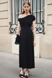 Model standing outdoors wearing an off-the-shoulder black dress with white stud details and holding a black handbag