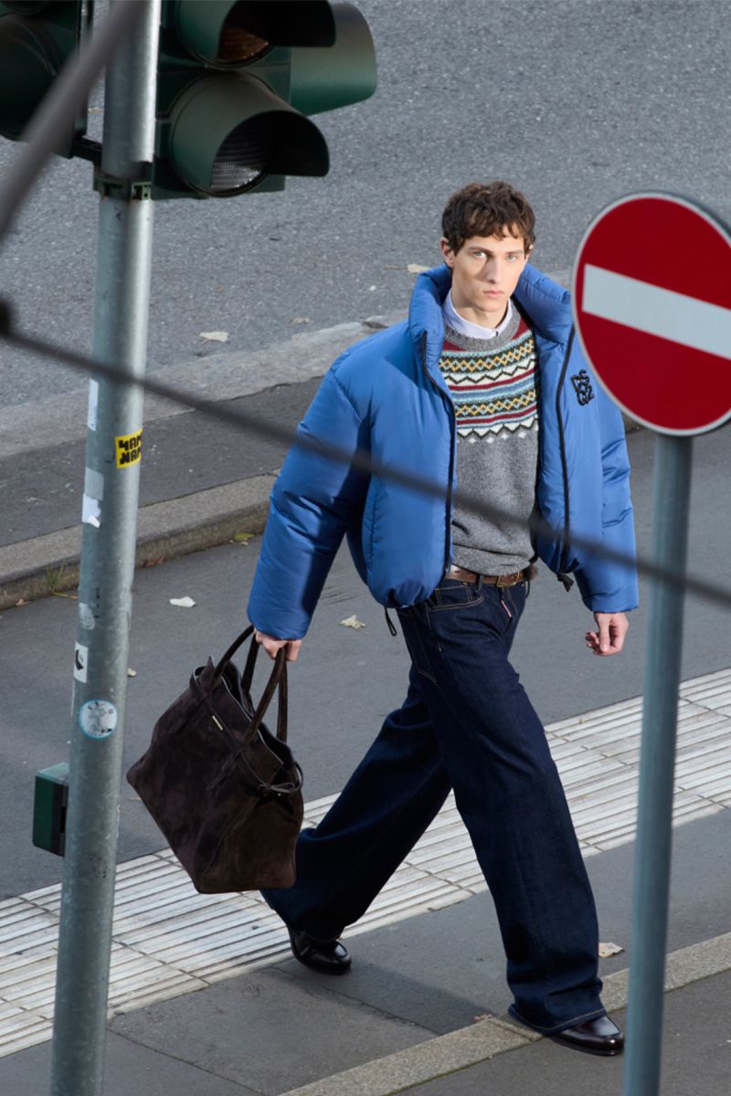 An adult man walks with a brown tote bag on a city sidewalk, wearing a blue puffer jacket and dark jeans; traffic lights and signs are visible.
