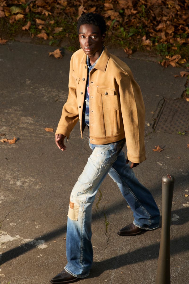 A young adult male in a tan jacket and distressed jeans is walking on a city street with leaves in the background during daytime.